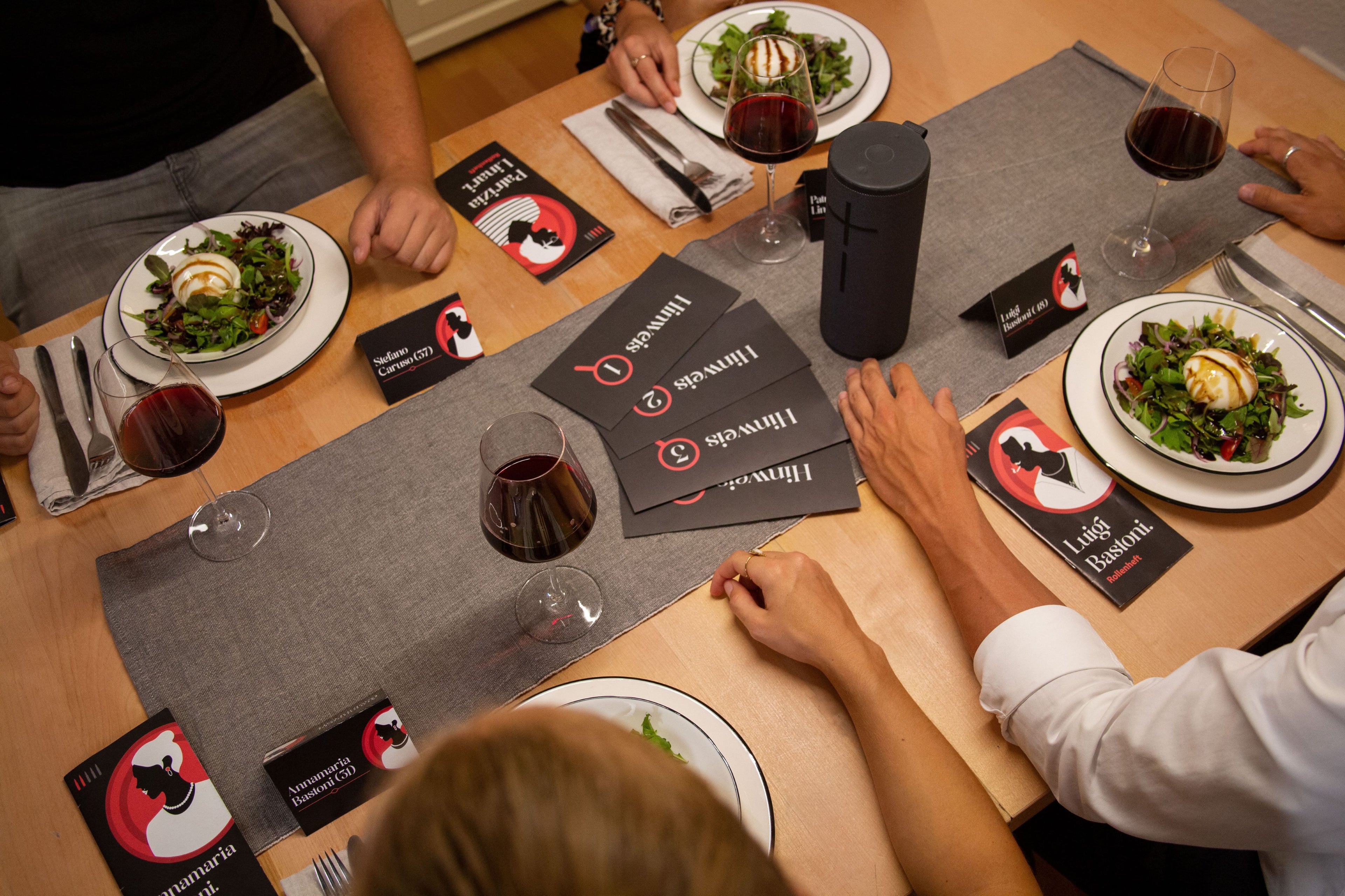 Ein für vier Personen gedeckter Tisch mit Salaten, Rotweingläsern und einem dunkelgrauen Becher. Schwarz-rote Placeit-Werbematerialien, darunter Untersetzer und Broschüren, liegen auf einem grauen Tischläufer. Vier Personen sitzen um den Tisch herum.