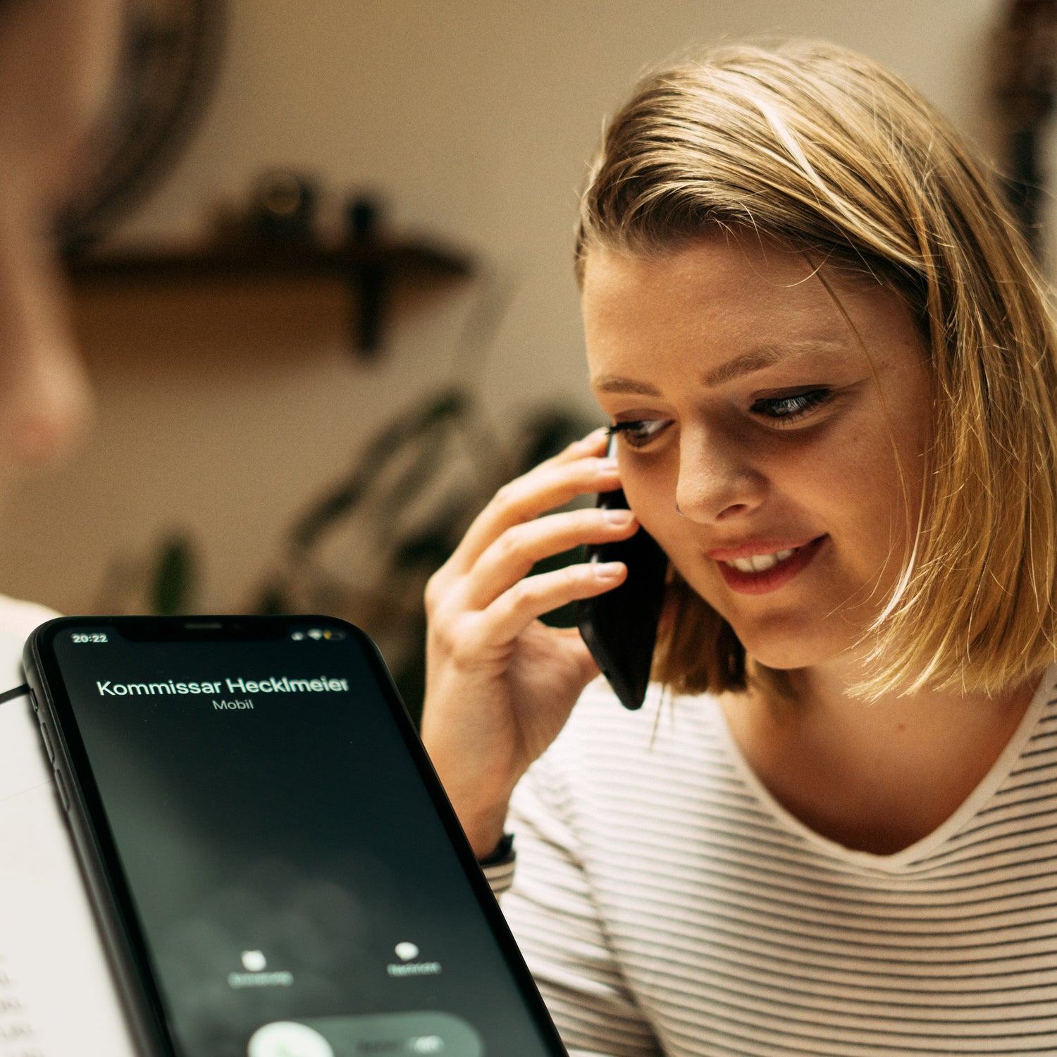 Eine blonde Frau hält lächelnd ein Telefon ans Ohr. Vor ihr zeigt das Display einen eingehenden Anruf von „Kommissar Hedimel“. Die Szene scheint in einem Raum mit Pflanzen im Hintergrund zu spielen.