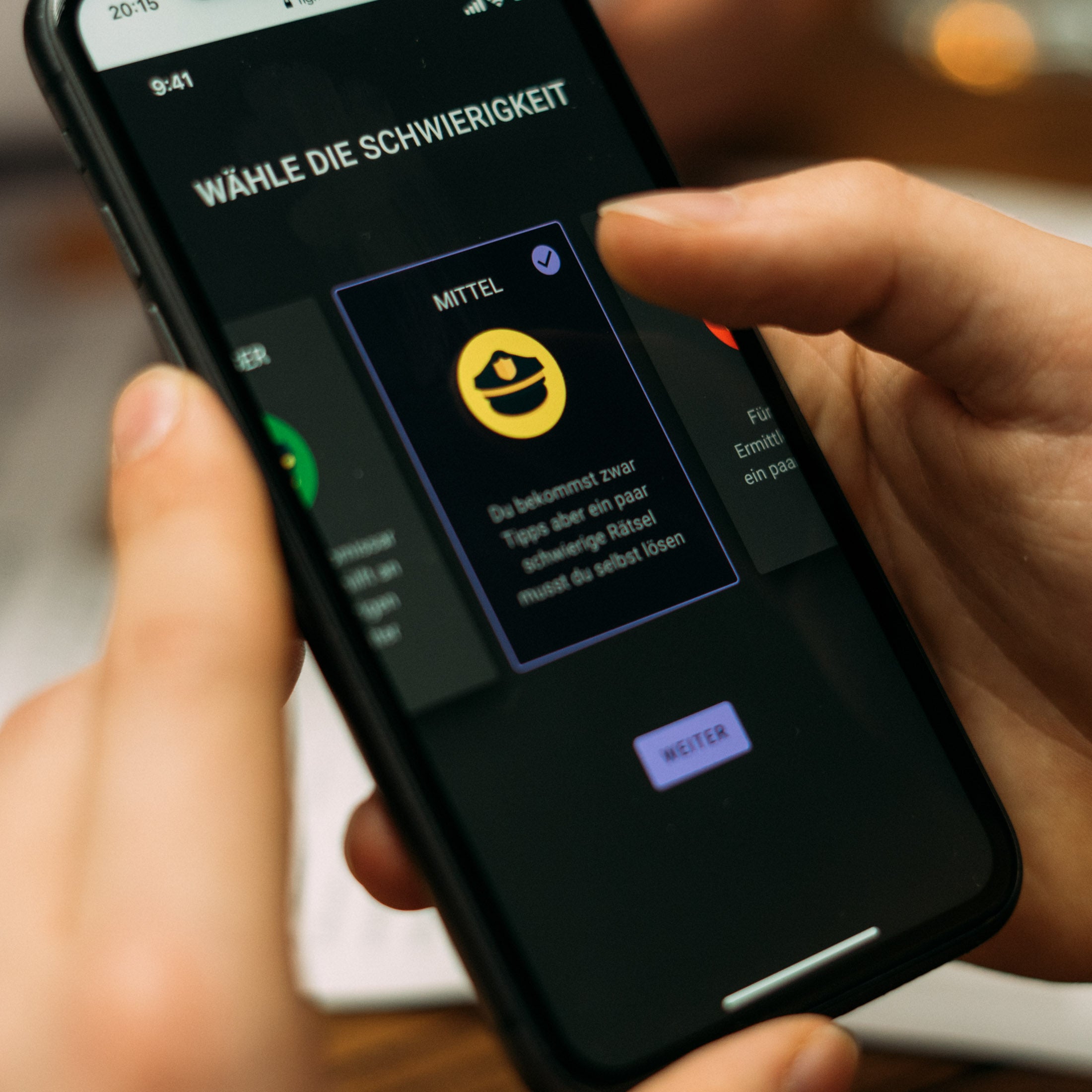 Eine Person hält ein Smartphone in der Hand, auf dem ein Menü zur Auswahl des Schwierigkeitsgrades auf Deutsch angezeigt wird. Die hervorgehobene Option zeigt ein gelbes Emoji mit Hut und der Aufschrift „Mittel“. Der Hintergrund ist unscharf.