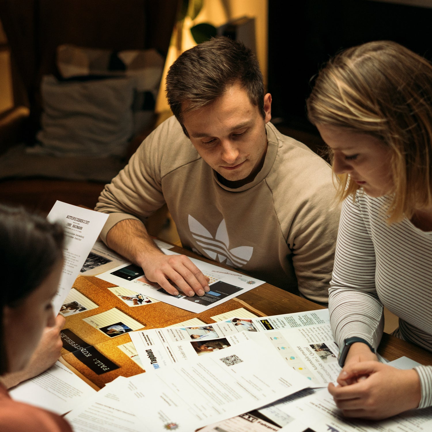 Drei Personen sitzen an einem mit Papieren und Dokumenten bedeckten Tisch, diskutieren und prüfen deren Inhalt. Eine Person trägt einen leichten Pullover mit Muster, die anderen sind leger gekleidet. Die Atmosphäre wirkt informell und gemütlich.