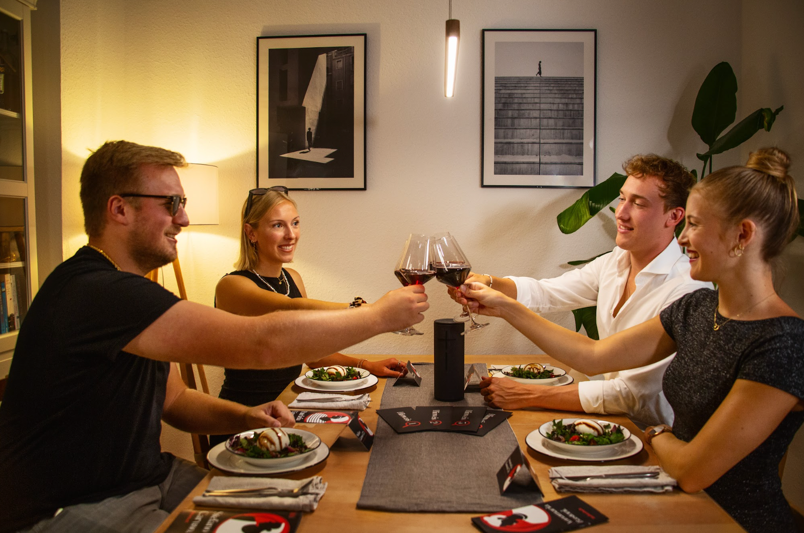 Vier Personen sitzen um einen Esstisch und stoßen mit Weingläsern an. Der Tisch ist mit Salaten und Menüs gedeckt. Zwei gerahmte Fotos hängen an der Wand, in der Ecke steht eine große Pflanze. Der Raum ist sanft beleuchtet.