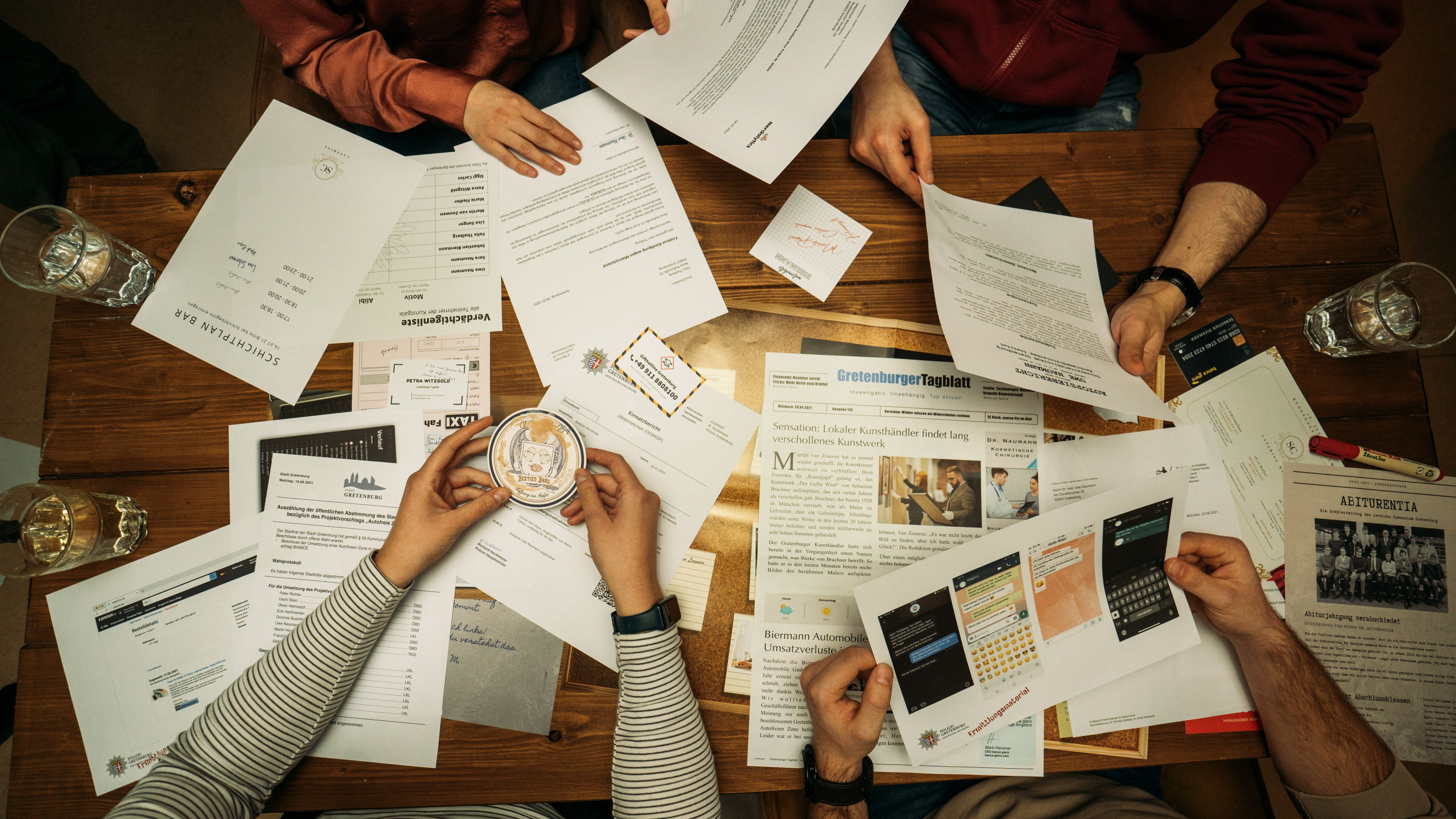 Ein Tisch ist mit verschiedenen Papieren und Dokumenten bedeckt, darunter eine Zeitung und ein Taschenrechnerausdruck. Vier Personen sitzen um den Tisch herum und sortieren und besprechen die Materialien. Es stehen auch Wassergläser bereit.