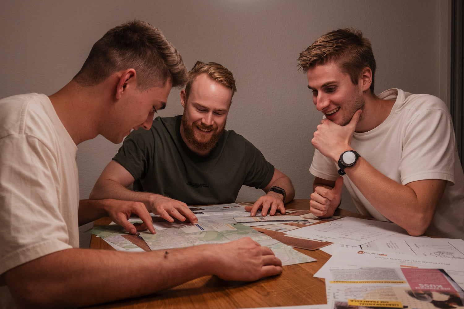 Drei Männer sitzen um einen Holztisch und prüfen Karten und Dokumente. Sie lächeln und scheinen etwas zu besprechen oder zu planen. Der Raum ist sanft beleuchtet, und vor ihnen liegen verschiedene Papiere ausgebreitet.