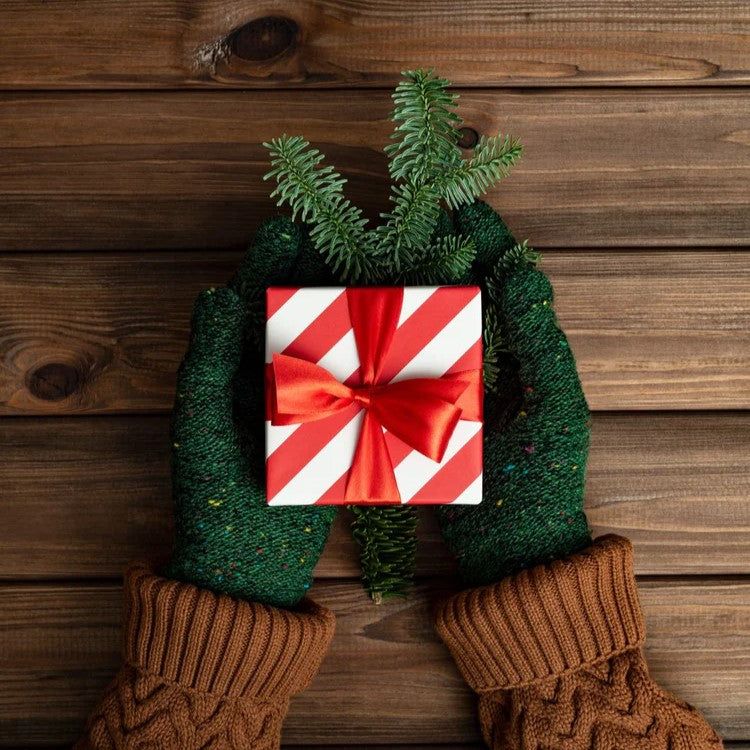 Grün behandschuhte Hände halten einen CRIMECASES-Geschenkgutschein, eine kleine, gestreifte Geschenkbox, die mit einer roten Schleife und Tannenzweigen verziert ist und auf einer Holzoberfläche liegt.