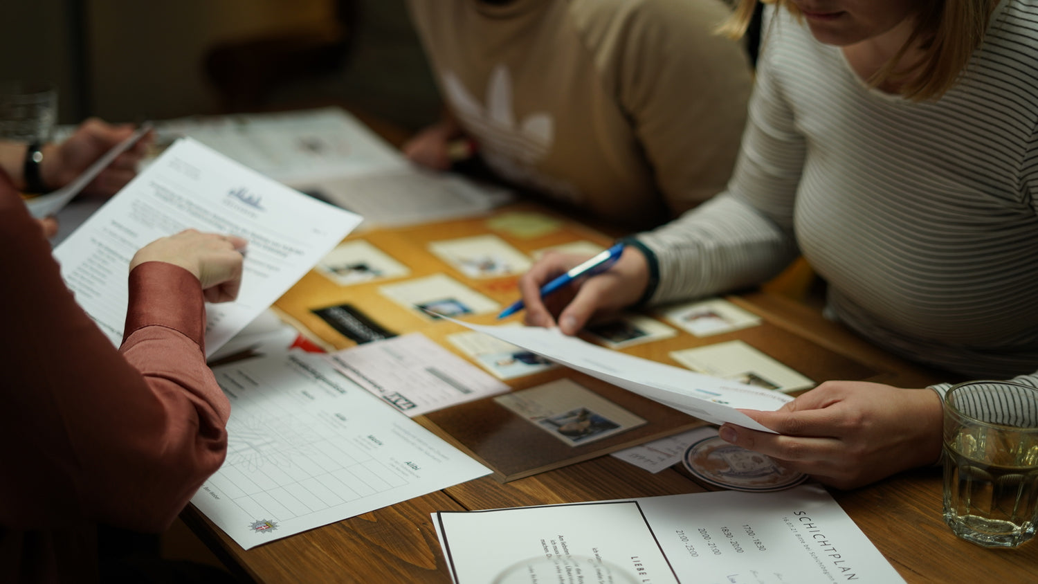 Drei Personen sitzen an einem Tisch und arbeiten an Dokumenten und kleinen Karten. Eine Person hält einen Stift und macht sich Notizen. Papiere und Karten mit Bildern und Text liegen ausgebreitet. Ein Glas Wasser steht daneben. Es scheint sich um eine Besprechung oder eine gemeinsame Aktivität zu handeln.