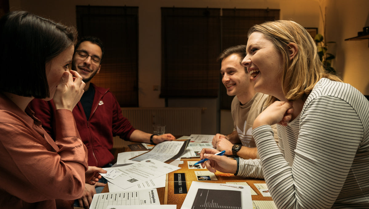 Vier Personen sitzen um einen mit Papieren und Büchern bedeckten Tisch. Sie unterhalten sich und scheinen zu lachen. Die Atmosphäre ist locker und entspannt, mit warmer Beleuchtung und einem Fenster mit geschlossenen Jalousien im Hintergrund.